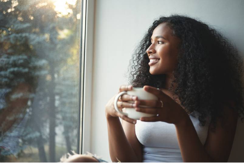 A young woman with curly black hair holds a coffee mug in both hands and looks out a window. She is smiling.