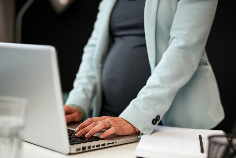 A pregnant person is standing at their desk typing on the laptop. They are wearing a dark grey shirt and a light blue blazer.