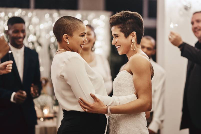 Two brides smiling and looking lovingly into each other's eyes as they embrace at their wedding reception.