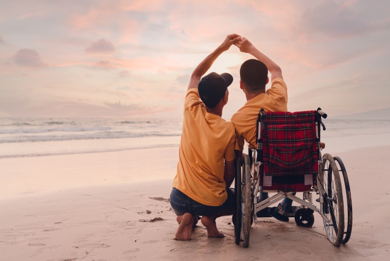 Two people are at the beach looking at the ocean water. One person is in a wheelchair, and the other squats next to them.