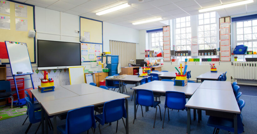 An empty elementary school classroom with large gray tables, blue chairs, posters, and learning tools.