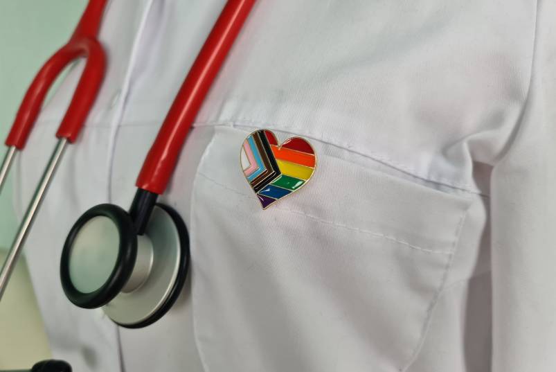 Close-up of doctor in a white lab coat with a red stethoscope around their neck wearing an LGBTQ+ pride pin.