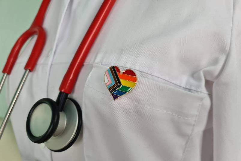 Close-up of doctor in a white lab coat with a red stethoscope around their neck wearing an LGBTQ+ pride pin.