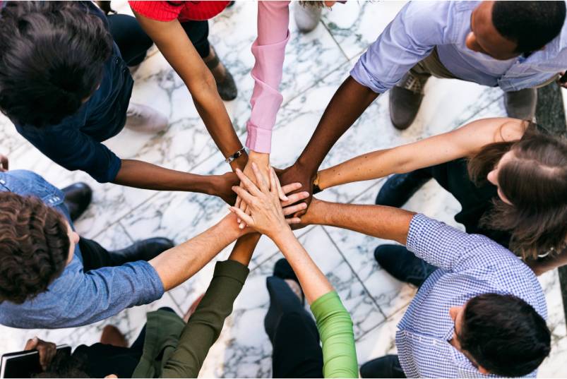 A group of nine people standing in a circle with their arms outstretched and hands stacked together in the center.