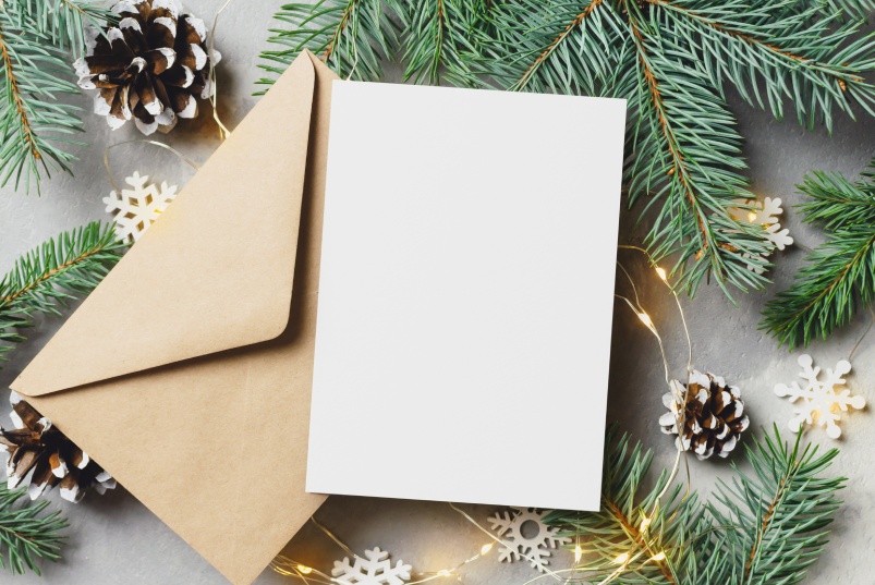 Closeup of a blank holiday card and envelope surrounded by pine tree branches, pinecones, and snowflakes.