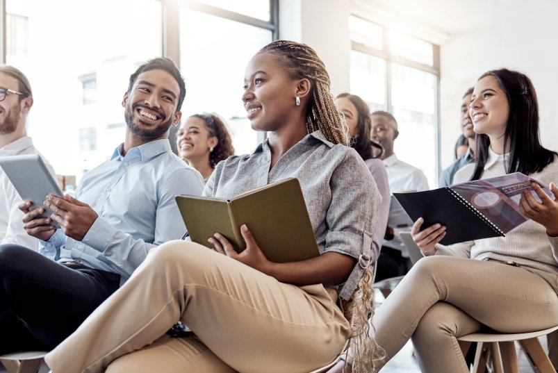 A diverse group of people sit in chairs, holding notebooks. They're all smiling and looking at the front of the room.