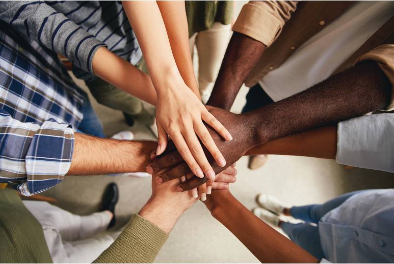 A group of people with various skin tones standing in a circle, each putting a hand forward in a team gesture.