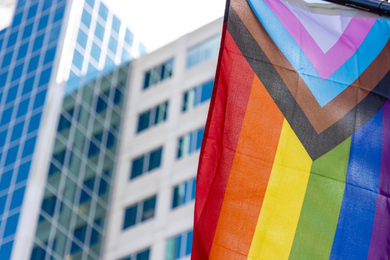 A Progress Pride flag flying in an outdoor area with skyscrapers visible in the background.