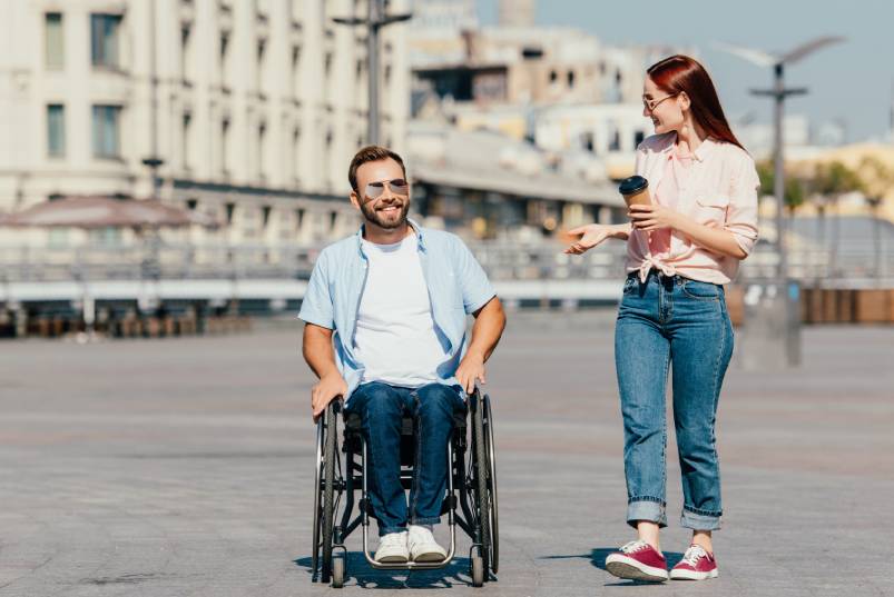 A man in a wheelchair and a woman holding a coffee cup walk together while talking on a sunny pier.