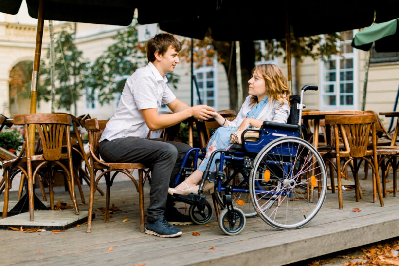 A man and a woman sit on an outdoor dining patio and hold hands. The woman sits in a blue wheelchair.