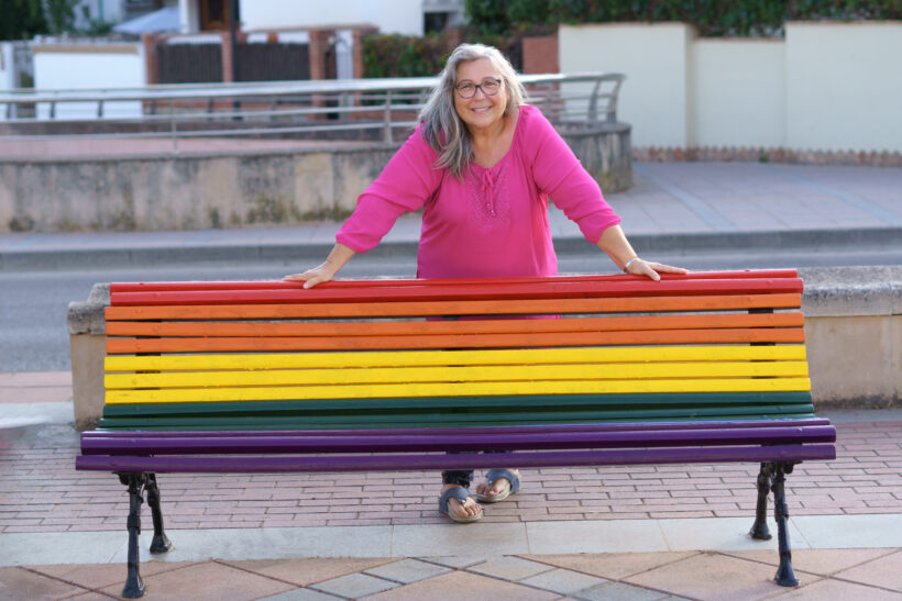 An older woman in a bright pink shirt leaning onto a bench. The bench rails are painted to look like a rainbow.