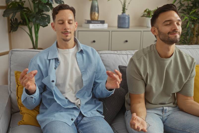 Two young men are sitting on a couch together with their eyes closed. They each have their hands out in a meditative pose.