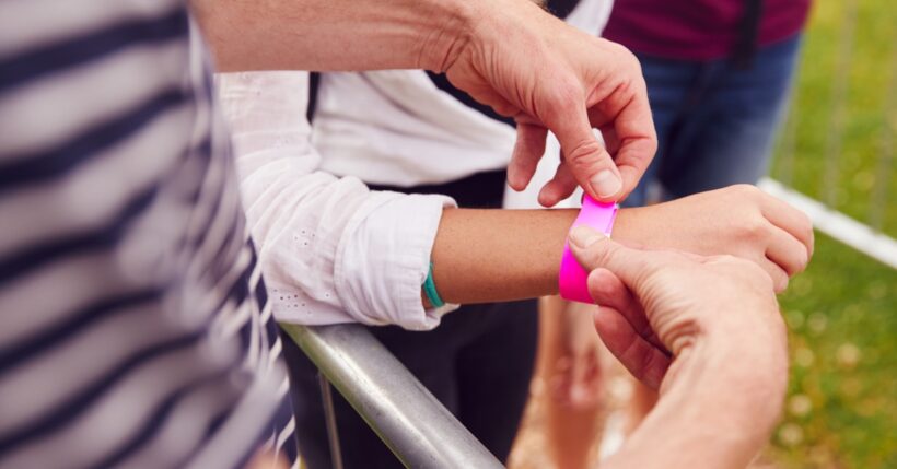 A close-up of a person at the entrance to an outdoor event putting a pink wristband on the wrist of another person.