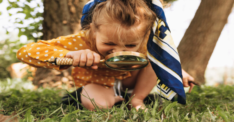 Outdoors, a little girl in a polka-dot dress holds a large magnifying glass. She crouches and uses it to look at the grass.