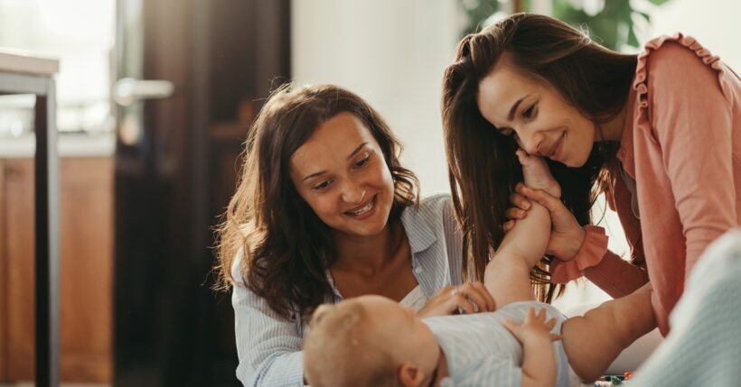 A WLW couple kneel in front of their baby, grinning affectionately at them. One mom holds the baby's foot against her cheek.