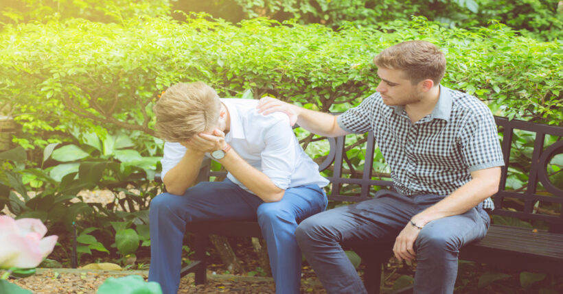 A person reaching over to hold the shoulder of their distressed friend. Both sit on a black bench in a garden area.