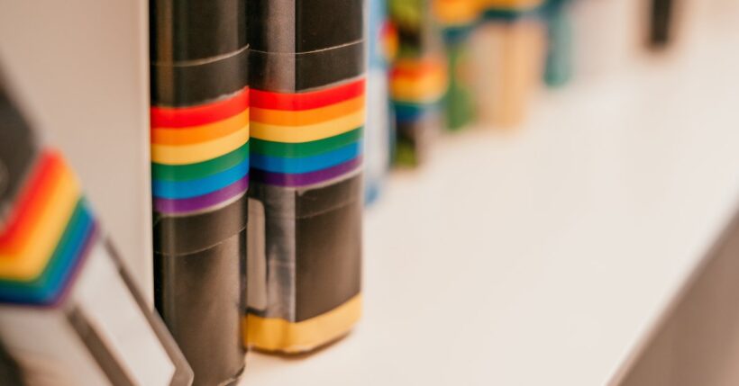 A close-up view of the spines of several books. Each one has a pride flag taped onto the lower half.