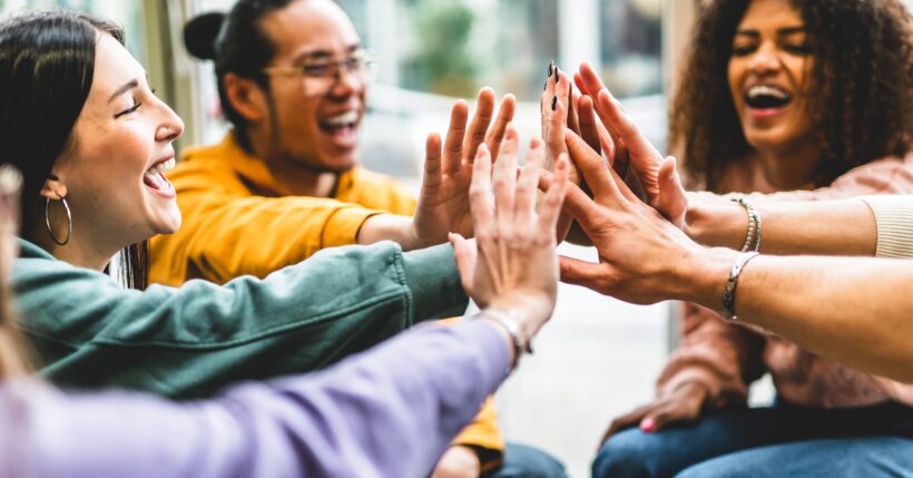 A diverse group of people wearing bright-colored clothes smiles happily and raises their hands together.