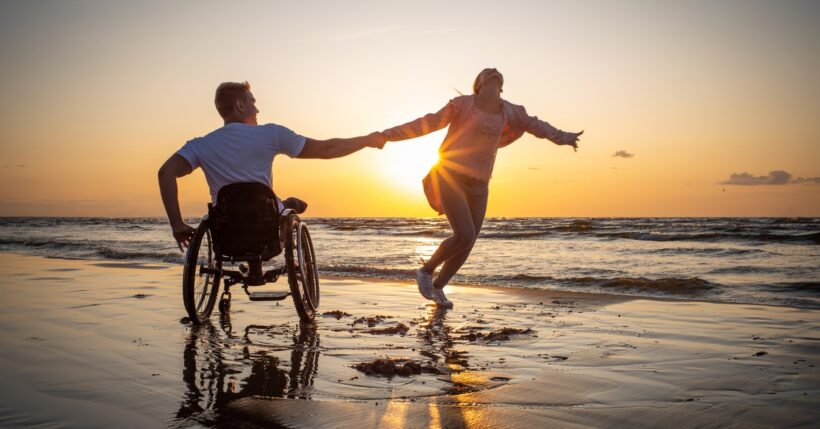 A handicapped man and an able-bodied woman moving along a beach. The sun is setting on the horizon in front of them.