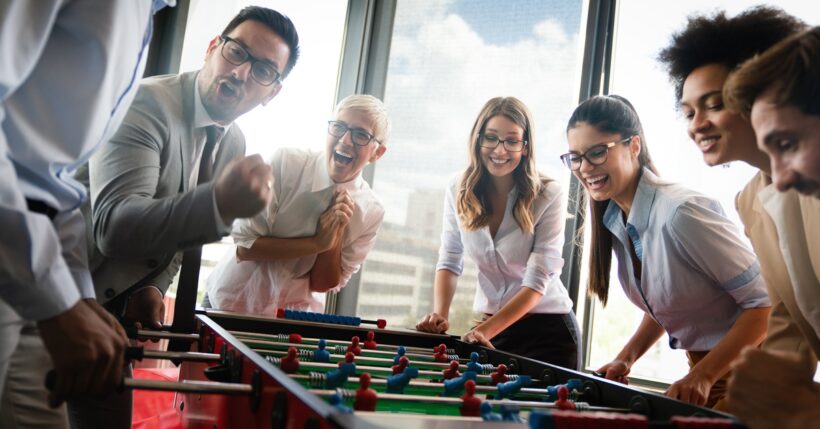 A diverse group of coworkers of all ages play foosball in an office, laughing and smiling in a lively, inclusive setting.