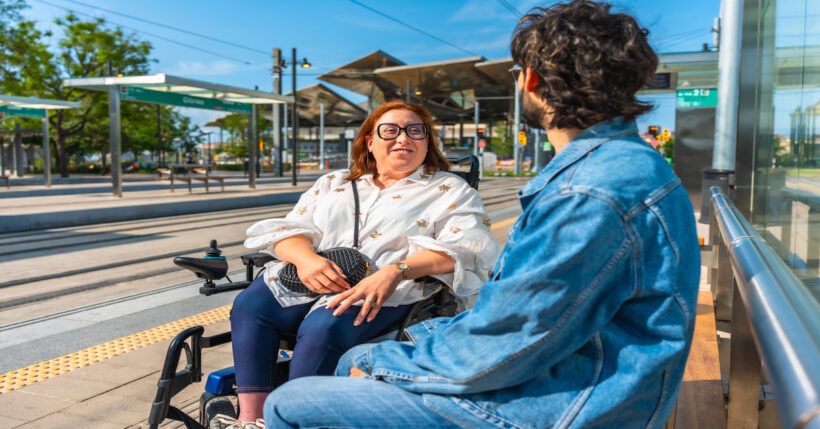 A woman sits in a powered wheelchair next to a tram stop. She smiles at a man sitting on the bench next to her.