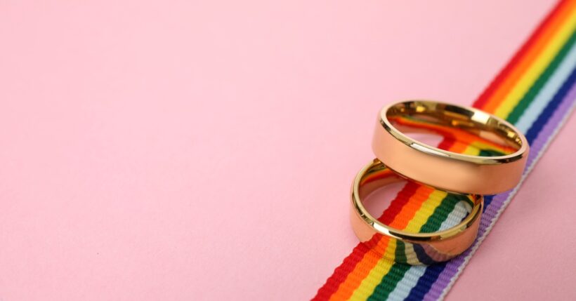 Two shiny gold wedding bands laid on a strip of rainbow ribbon against a solid pink background.