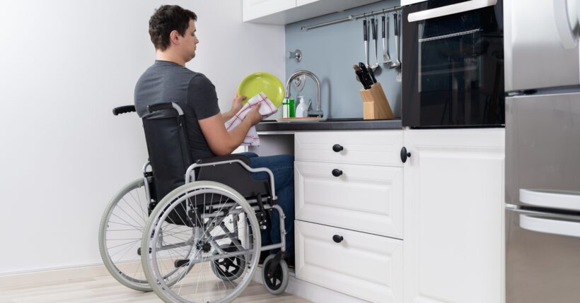 A man in a wheelchair doing dishes at an accessible kitchen sink. He dries a yellow dish with a white and red towel.