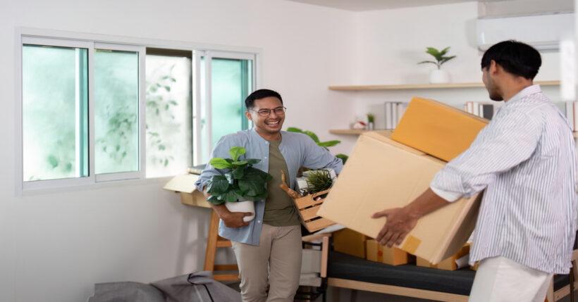 A gay couple moving in together. One guy is laughing while holding plants, and the other carries two boxes into the room.