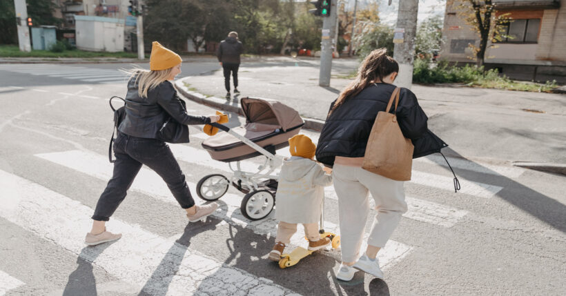 Two women and their children are crossing a crosswalk. One of the women pushes a baby stroller while the other guides a child on a scooter.