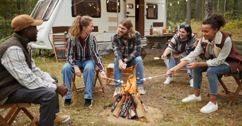 Five people are sitting in wooden chairs around a small campfire next to an RV. They're roasting marshmallows.