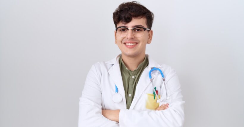 A smiling young LGBTQIA doctor in a white coat with a stethoscope and pens stands confidently with their arms crossed.