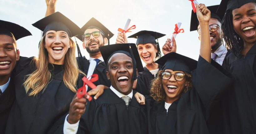 A group of racially diverse graduate students smiles and holds their diplomas in the air while outside.