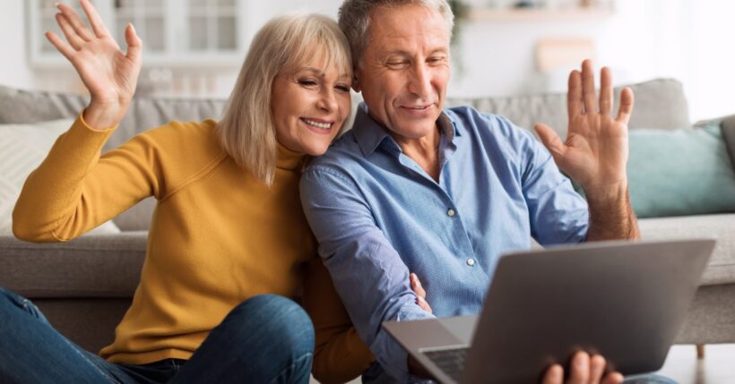A senior couple on a video call, smiling and waving at their laptop. They sit on the ground in front of their couch.