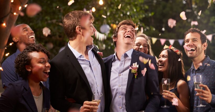 A group of men and women dressed in formal attire, smiling and holding champagne glasses while gathered outdoors.