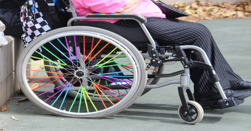 A wheelchair user outside on a sunny day. Their wheelchair spokes and armrests are rainbow colors, and they carry a backpack.