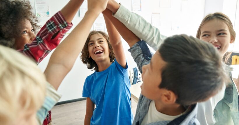 A close-up of five diverse kids huddled together in a brightly lit classroom, giving each other a high-five.
