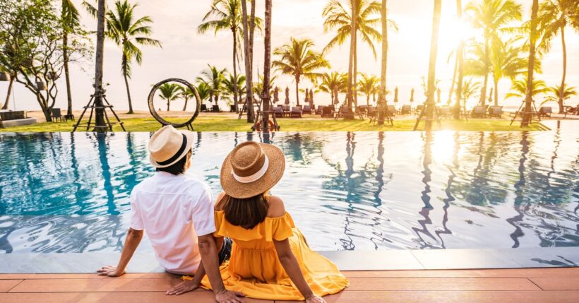 A young couple wearing straw hats lounge and sit by the pool watching the sunrise, surrounded by palm trees.