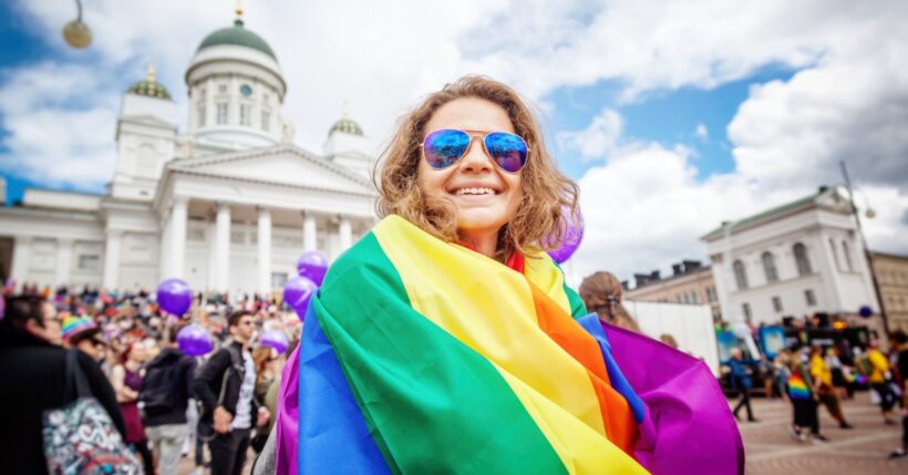 A young woman smiling and holding a rainbow flag while standing on a street in a European city with buildings behind her.