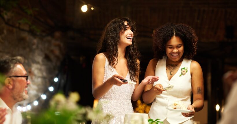 A pair of brides in white performing the cake cutting ceremony at a wedding while friends and family watch.