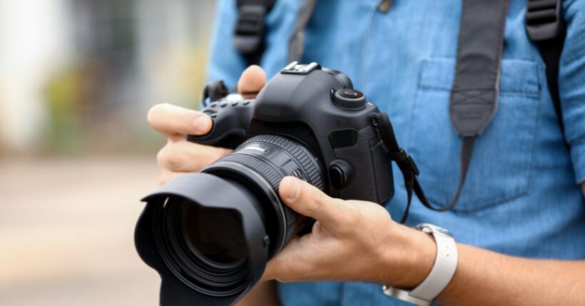 A person in a blue button down shirt holding a digital camera in their hands. There is a smart watch on their wrist.