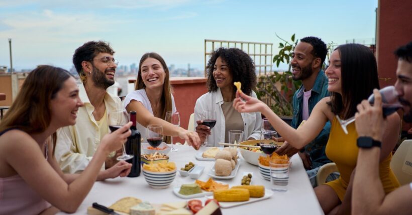 A diverse group of friends share food and wine while laughing together around a table on a sunny rooftop terrace.