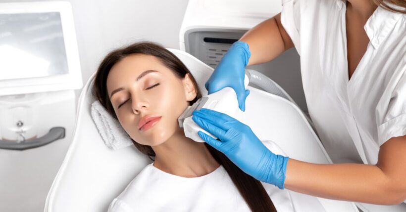 A young woman sitting in a medical chair as a woman with blue gloves uses a laser hair removal device on her jaw.