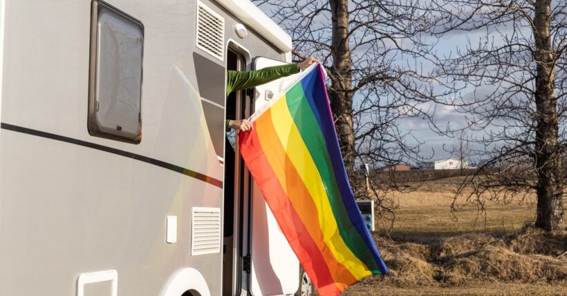 A rainbow flag is presented from a camper parked on a roadside with mountains and clouds in the sky behind it.