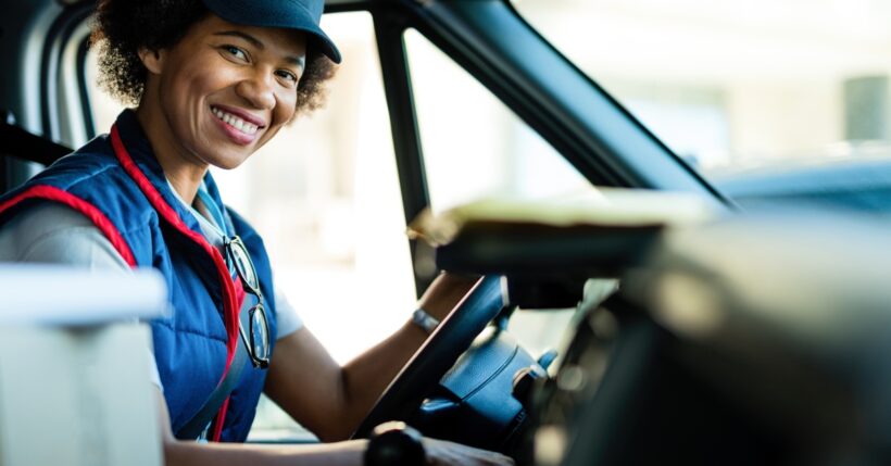 A smiling woman sitting in a van driver's seat with stacked boxes on the seat beside her and a hand on the steering wheel.