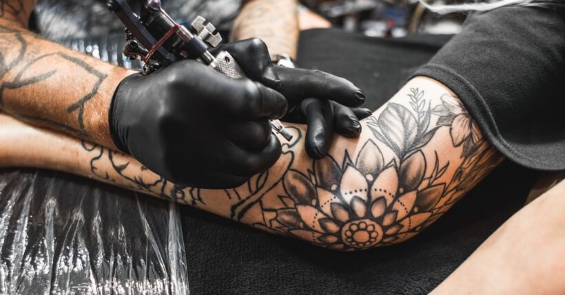 A tattoo artist using a tattoo gun to print a floral pattern tattoo onto the outstretched arm of a man.