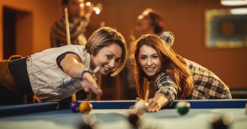 Two women playing pool or billiards in a pool hall with others having a conversation behind them as they play.