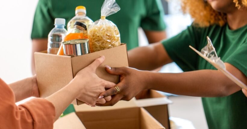 A volunteer in a green shirt and another person holding a cardboard box of bottled water and canned food.