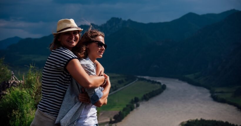 Two people embrace at a scenic overlook above a winding river, with dark mountains under stormy skies.