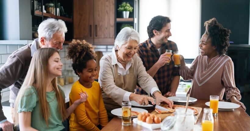 An extended family gathers around a kitchen table while preparing food together, smiling and sharing a relaxed moment.