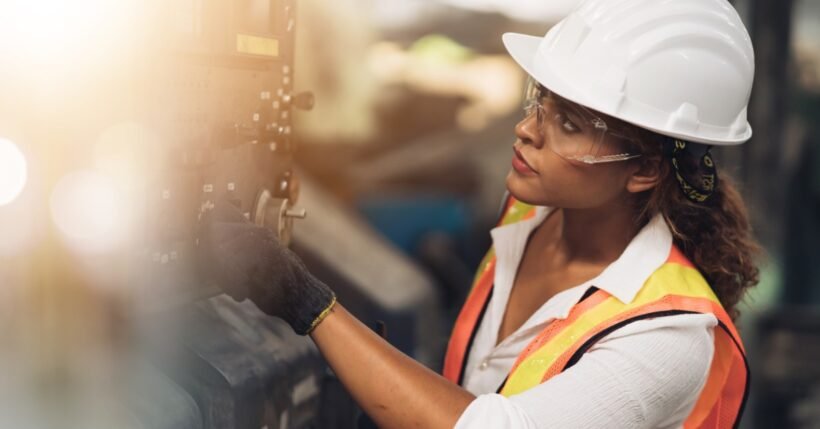 A female tradesperson in a hard hat and safety vest operates an industrial control panel on the shop floor.